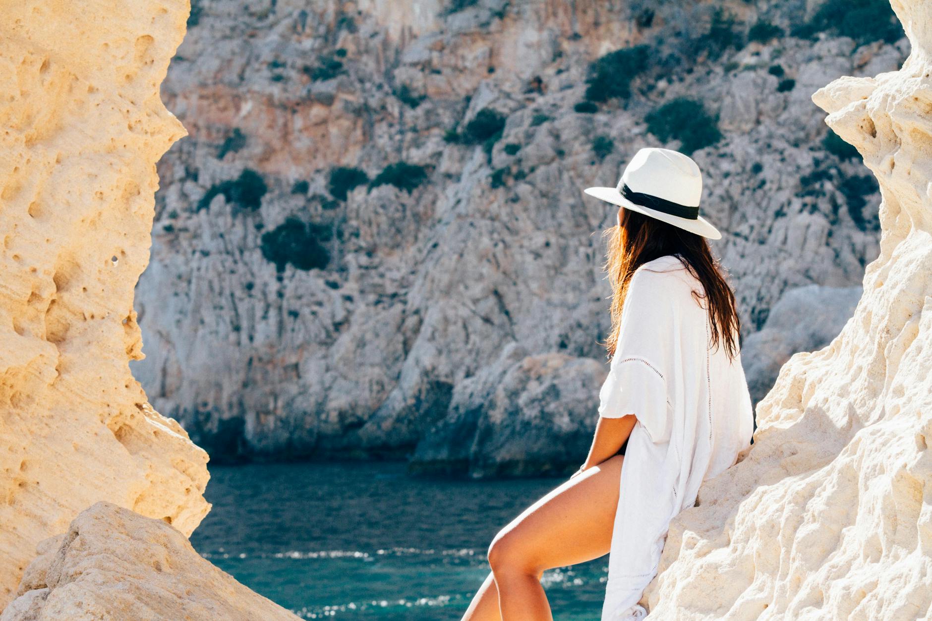 A woman in a hat sits on a rocky beach in summer, enjoying the sea view. [pexels]
