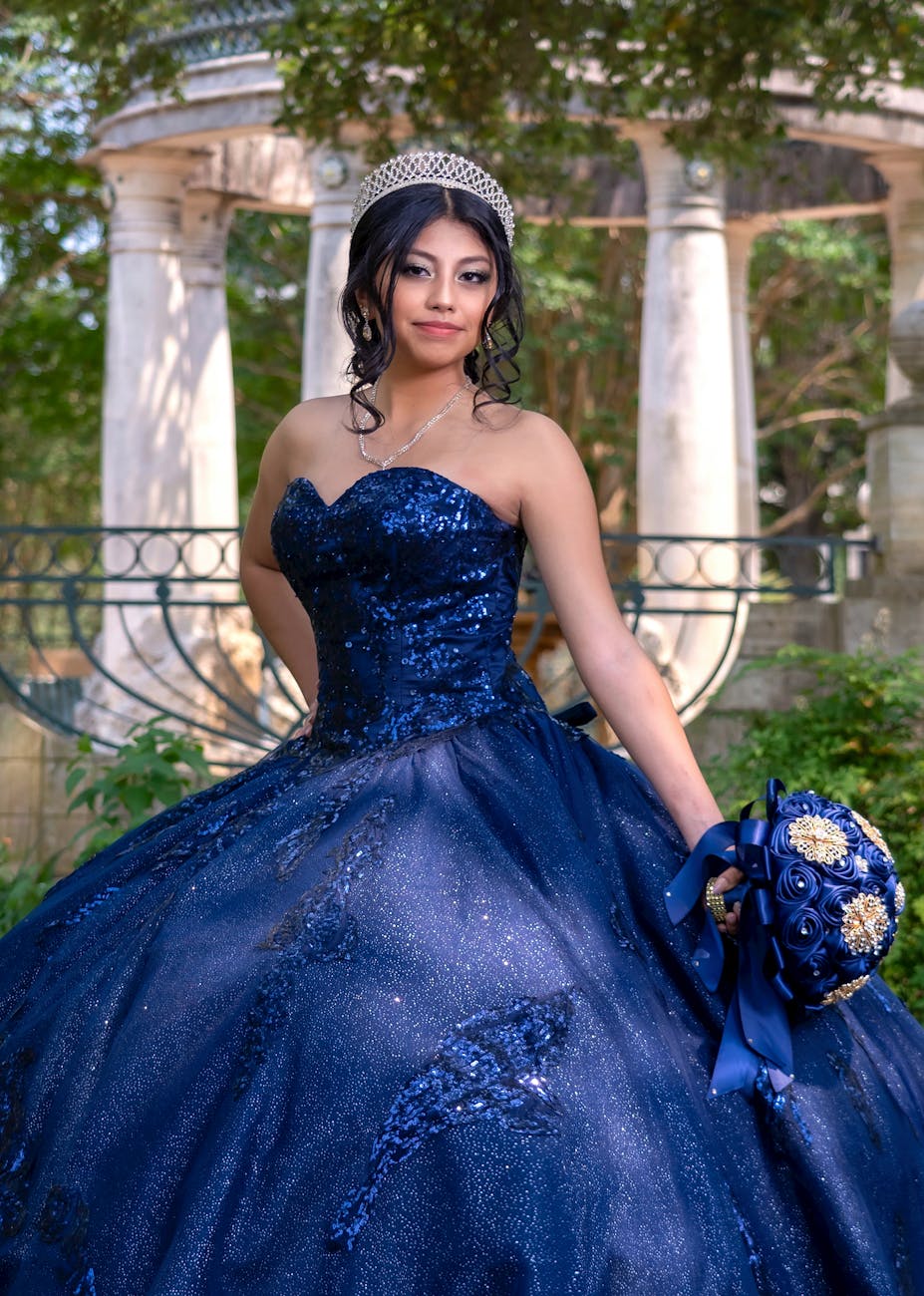 A young woman in a glittering blue quinceañera gown poses elegantly in an outdoor setting with grand columns. [pexels]