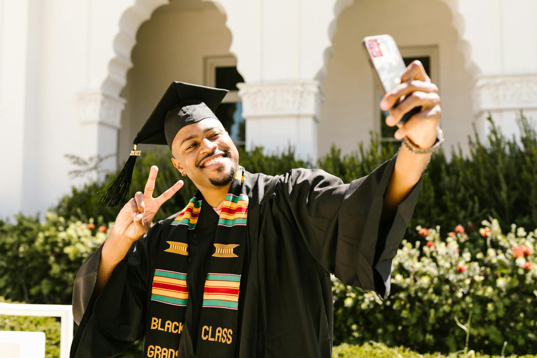 Joyful graduate in cap and gown taking a selfie outdoors for celebration. [pexels]