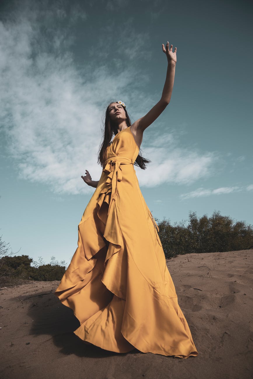 Stylish woman in a yellow dress posing dramatically on a sandy hill with a blue sky backdrop. [pexels]
