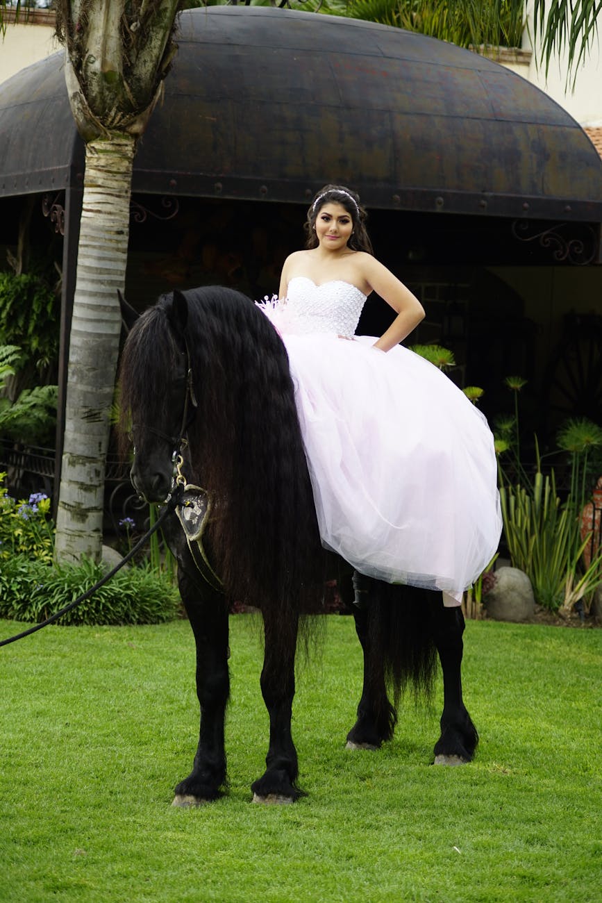 Young woman in a pink gown riding a black horse in a lush outdoor setting. [pexels]